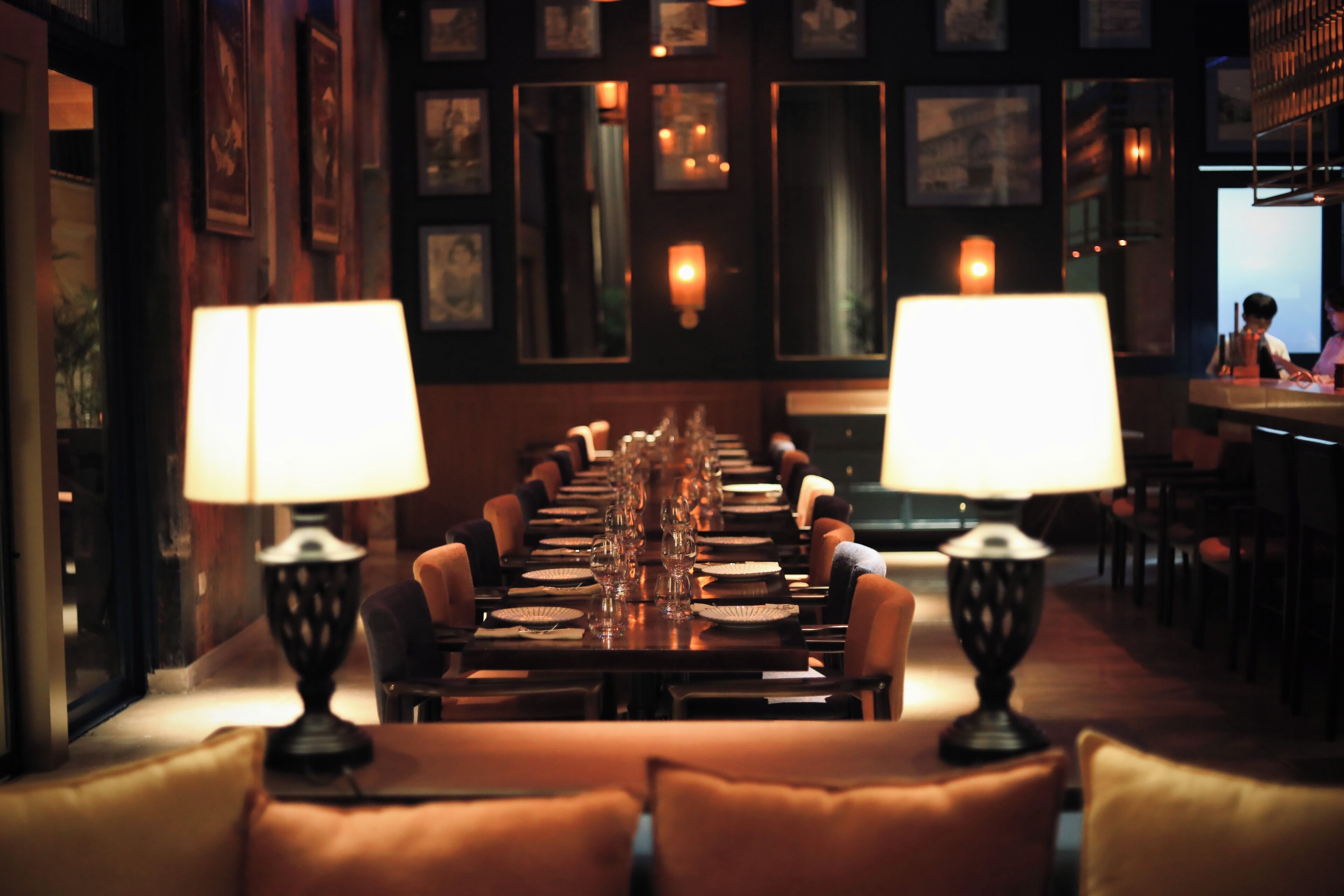 A long dining table with multiple chairs, lit by two lamps, in a dimly lit room with framed pictures on the walls.