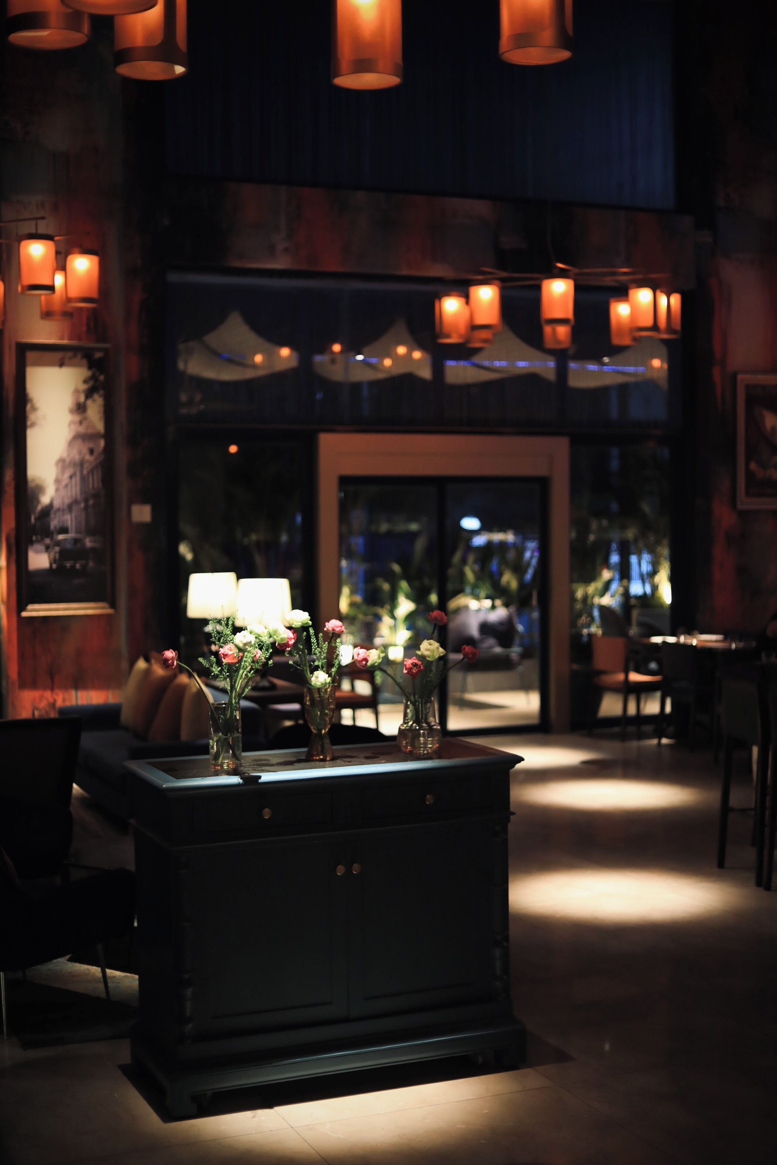 A dimly lit lobby with a central table displaying vases of flowers, a dark wooden cabinet, and hanging lanterns.