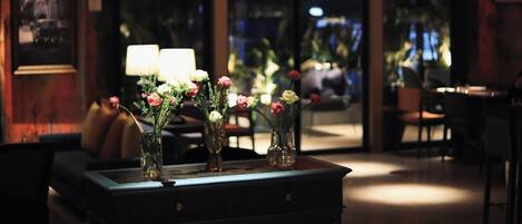 A dimly lit lobby with a central table displaying vases of flowers, a dark wooden cabinet, and hanging lanterns.