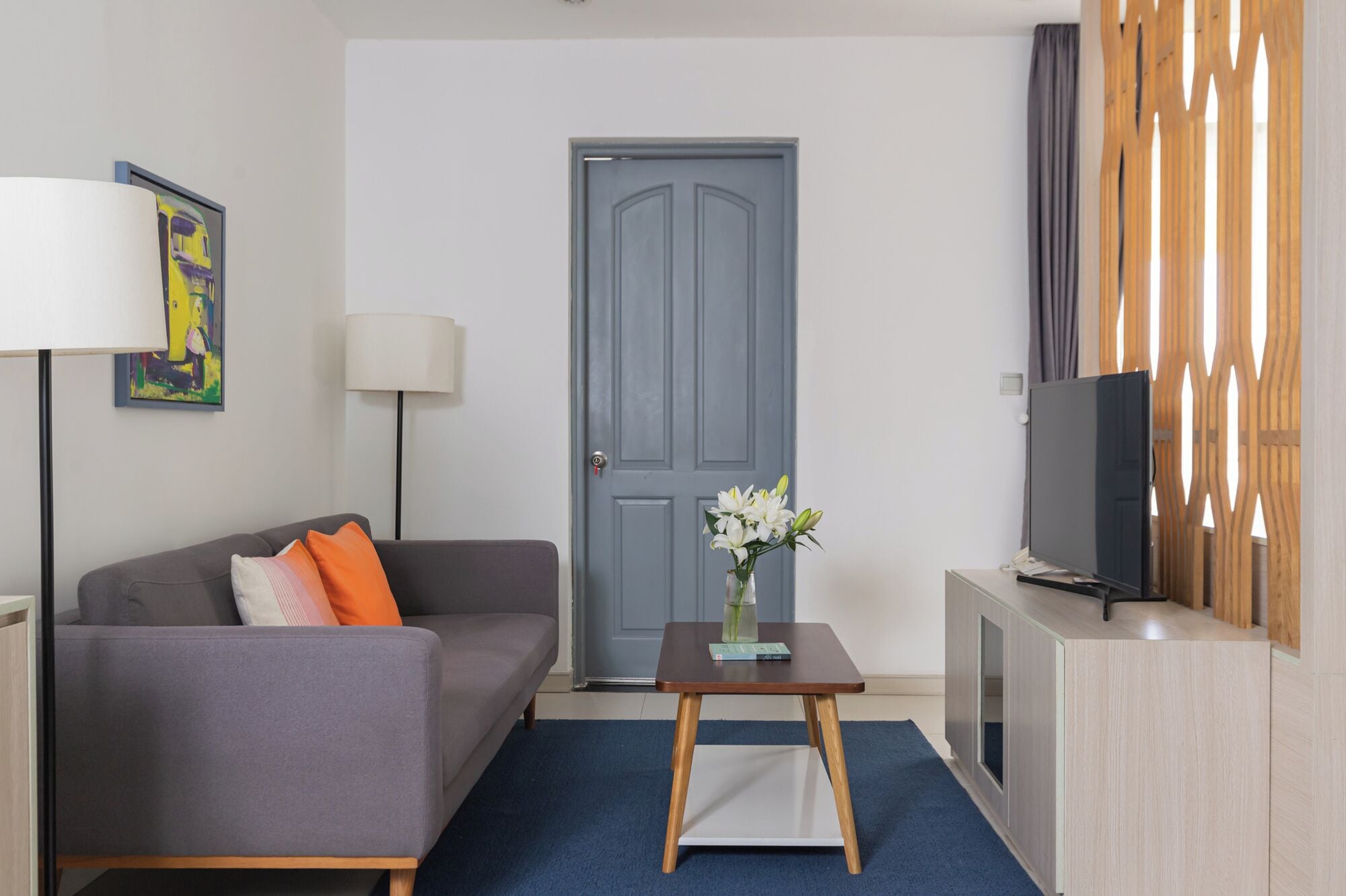 A modern hotel room with a grey sofa, a small table with a vase of flowers, a flat-screen TV on a wooden cabinet, and a large grey door.