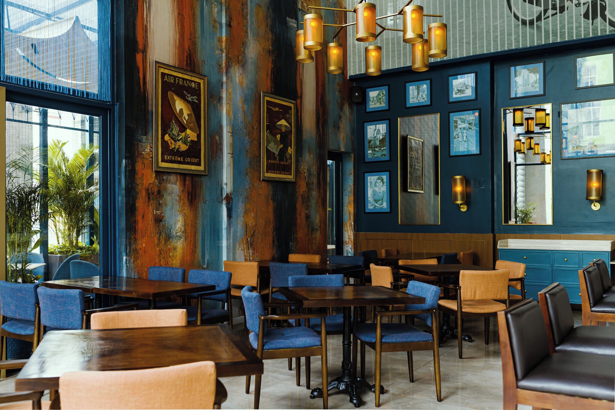 A restaurant interior with wooden tables and blue chairs, framed pictures on the walls, and a large window with a view of palm trees.
