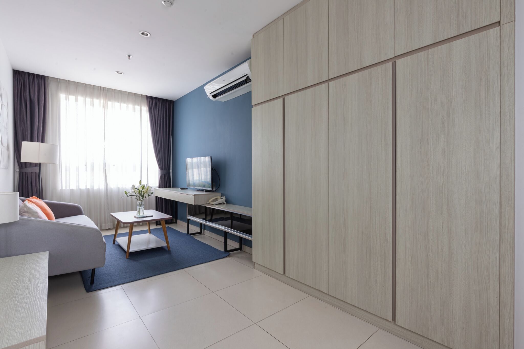A modern hotel room with a grey sofa, a small table with a vase, a desk with a computer, and a wardrobe.
