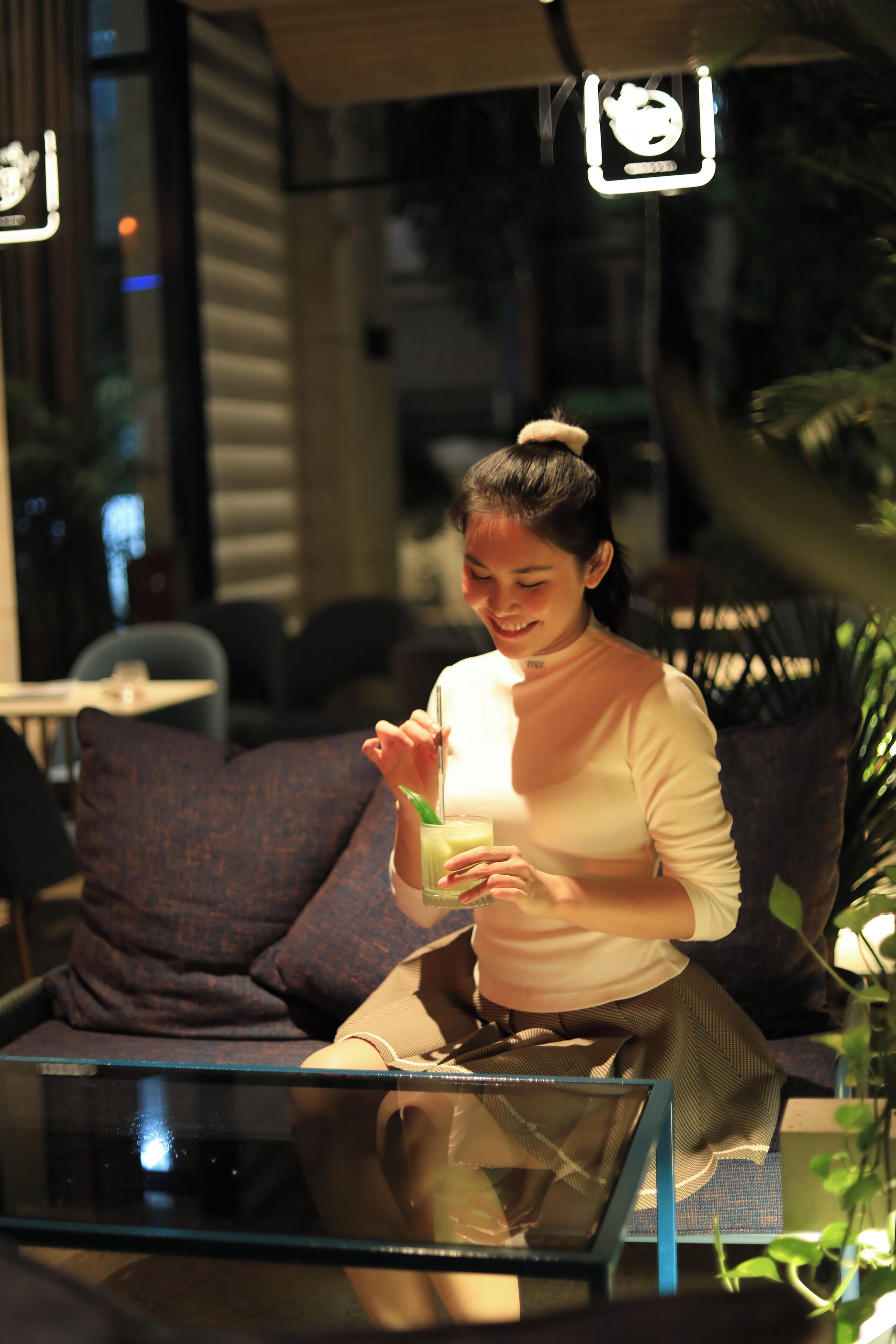 A woman sitting on a couch with a glass of drink, in a well-lit indoor setting with modern furniture and plants.