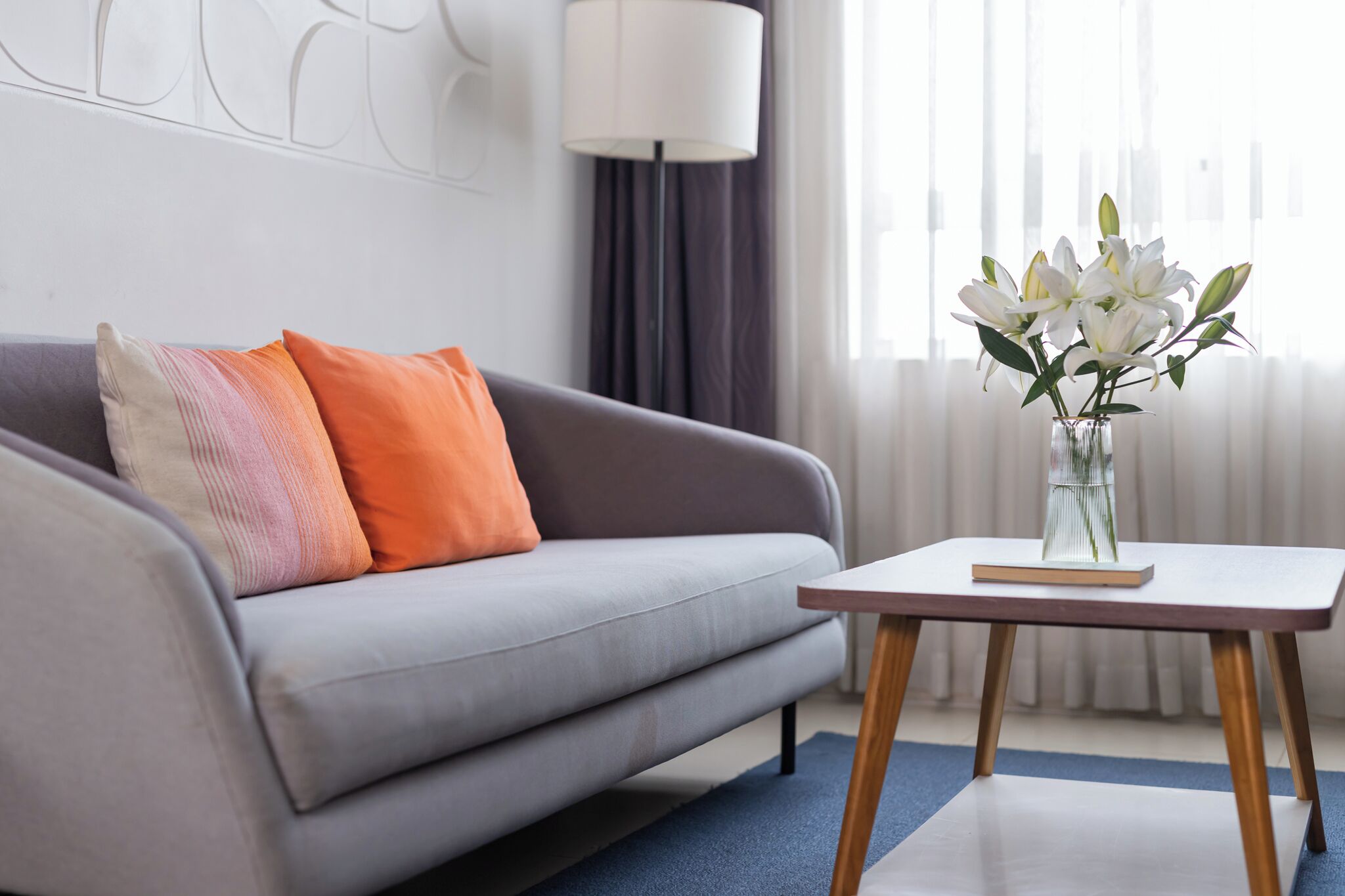 Una sala de estar moderna con un sofá grisáceo, cojines naranja y rosa, un jarrón blanco con flores y una pequeña mesa de madera.