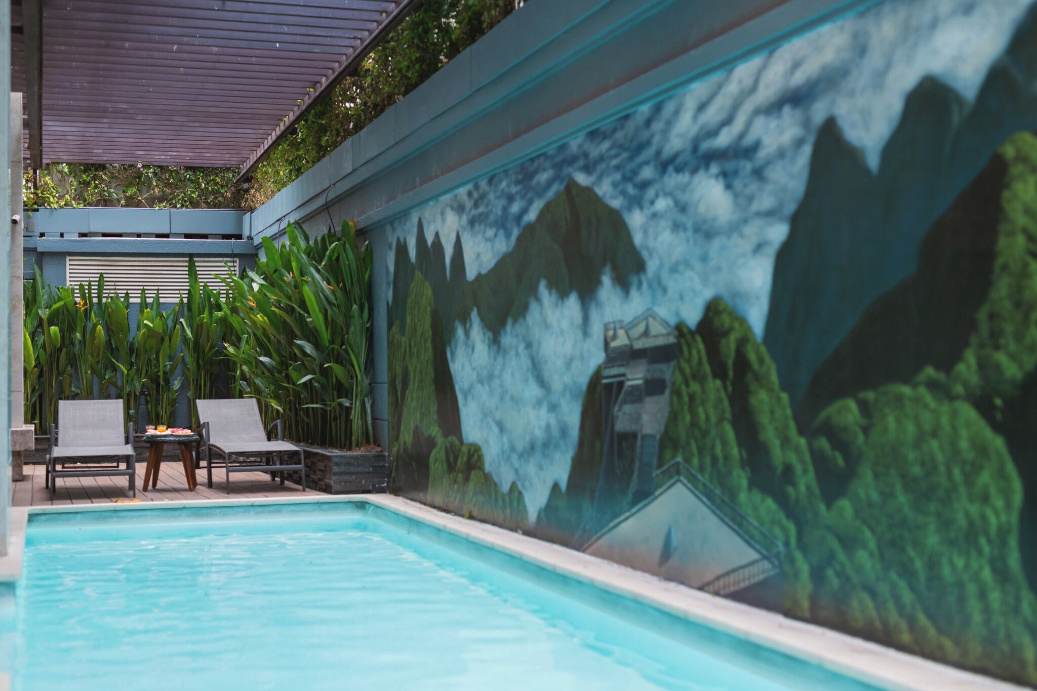 A swimming pool with a mural of mountains and a covered area with lounge chairs and a table.
