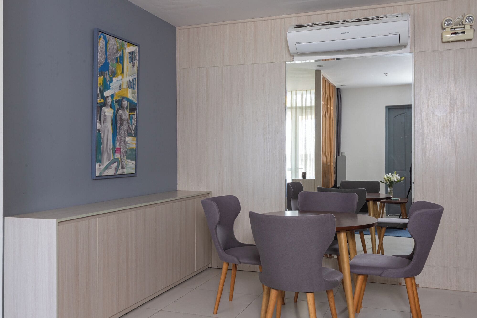 A modern dining area with a gray wall, a painting, and a large mirror reflecting the room.