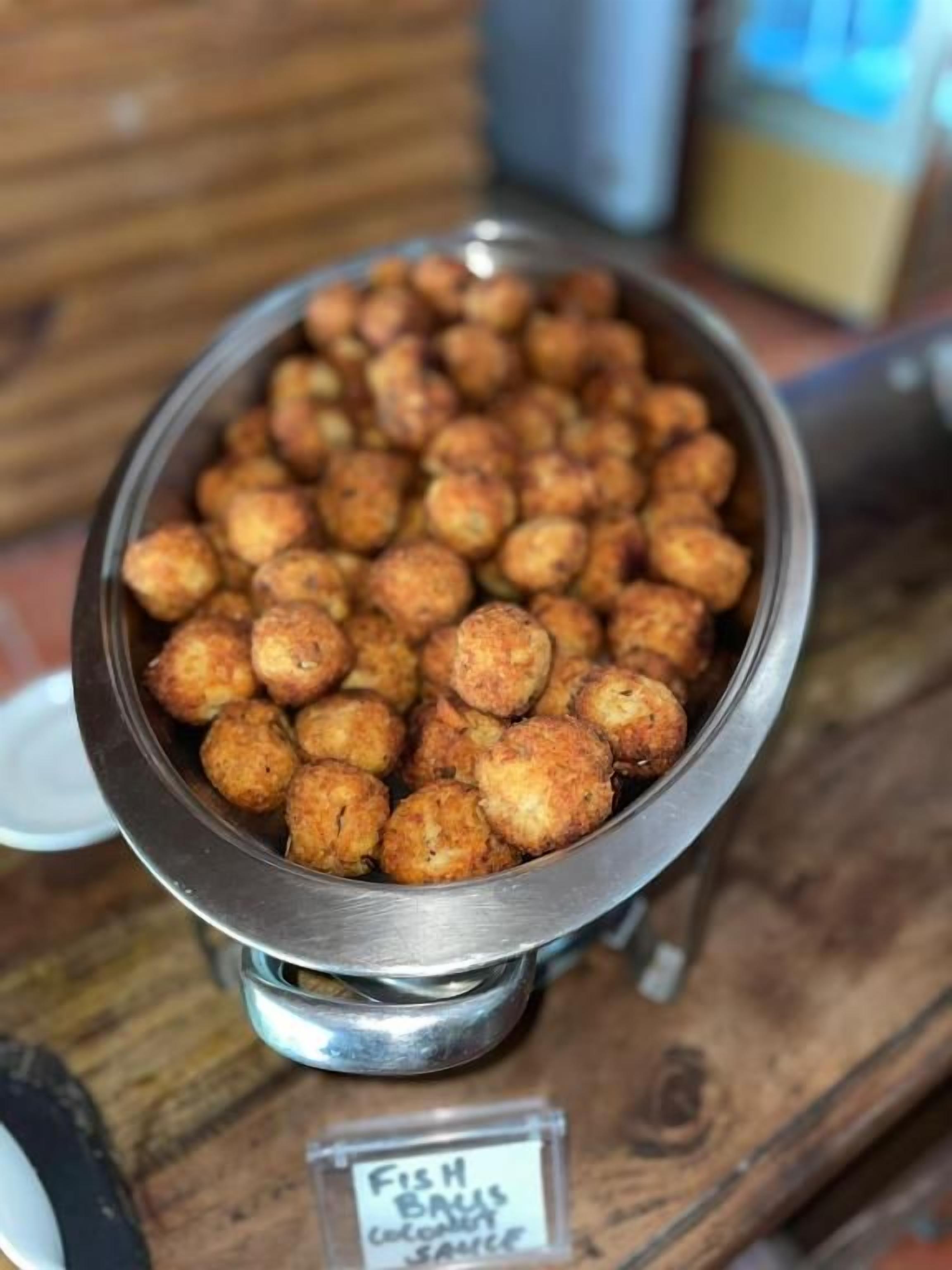 A metal bowl filled with fried food items, possibly fritters, with a label indicating 'Fish Balls' and 'Coconut Sauce' in the background.