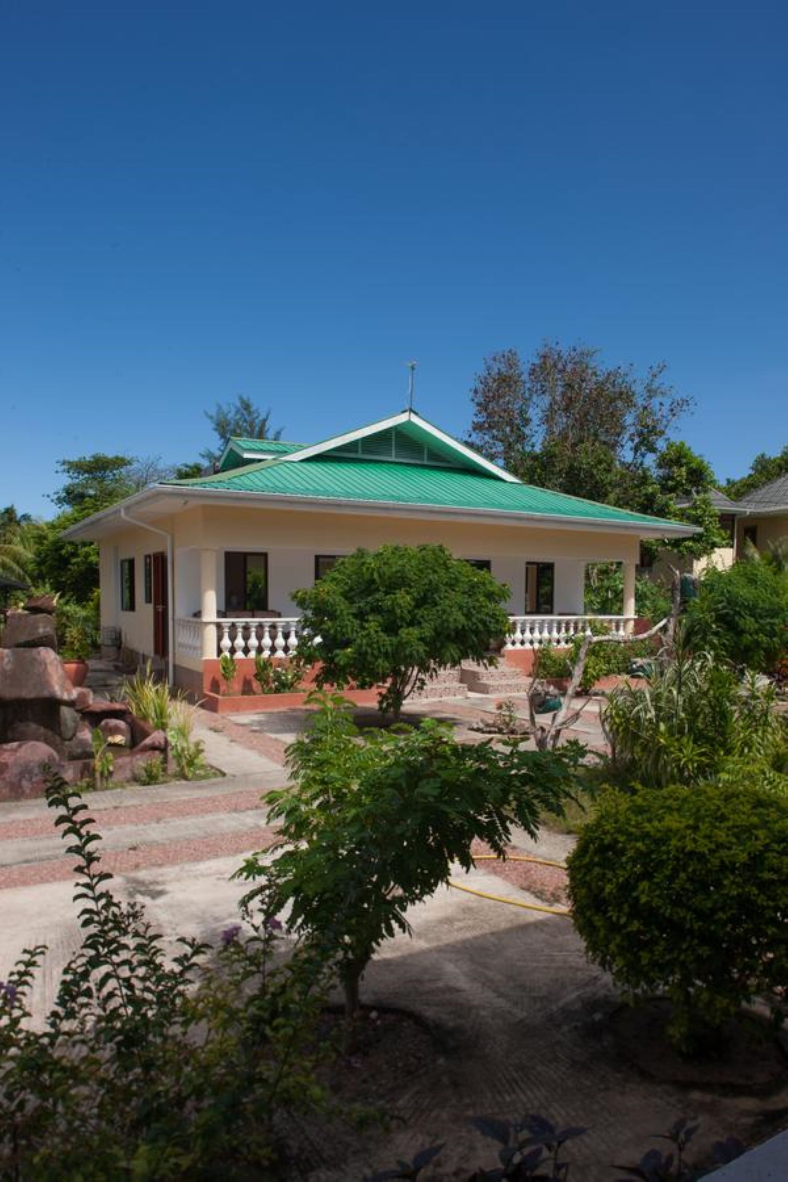 Uma casa térrea com telhado verde, varanda com guarda-corpo branco e jardim com diversas plantas e uma pequena árvore.