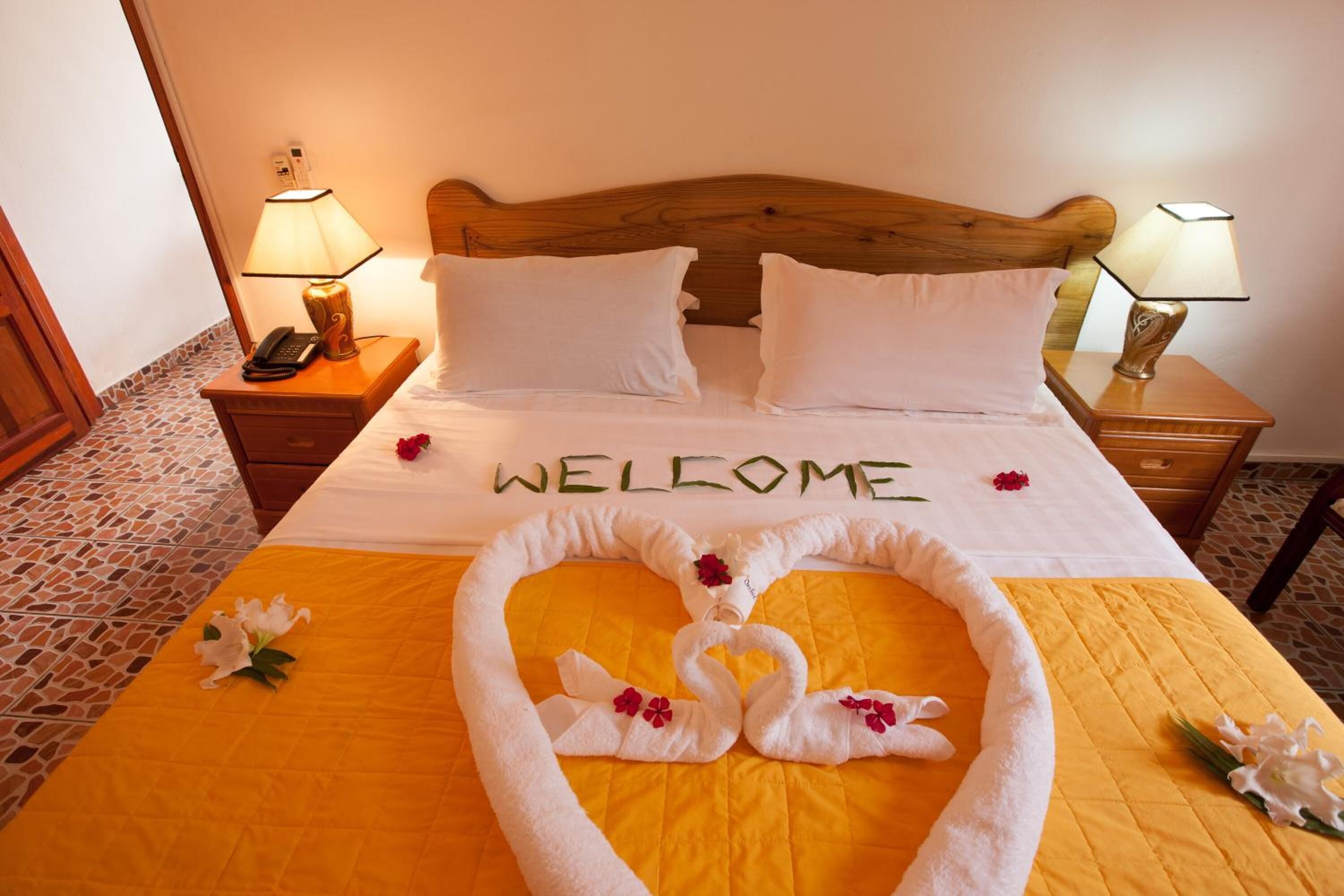 A neatly made bed with a 'WELCOME' sign, a heart-shaped towel arrangement, and a 'WELCOME' sign on the wall.