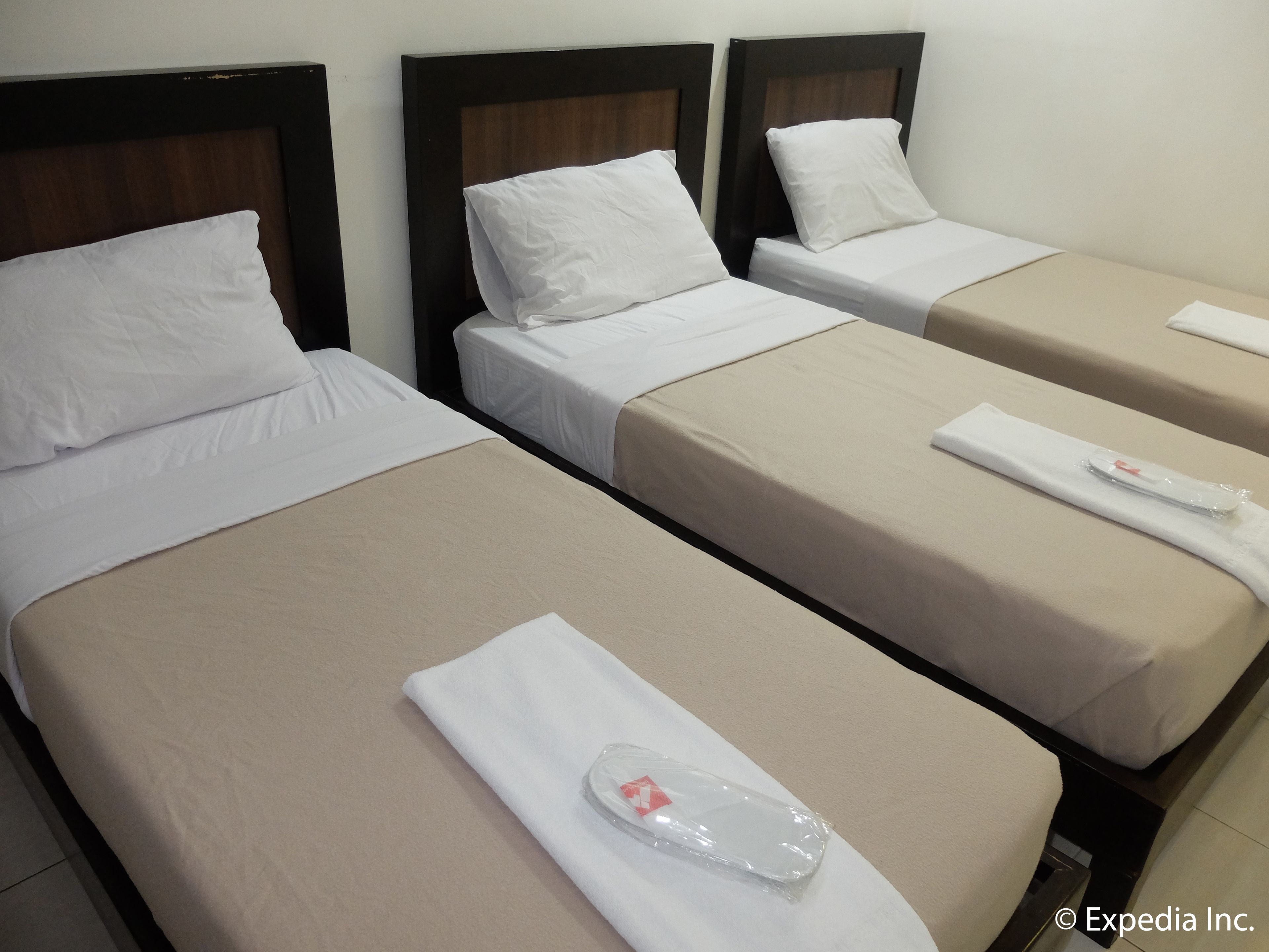 Three single beds with wooden headboards, white linens, and beige bedspreads. Each bed has a white pillow and a neatly folded towel with a small red tag.