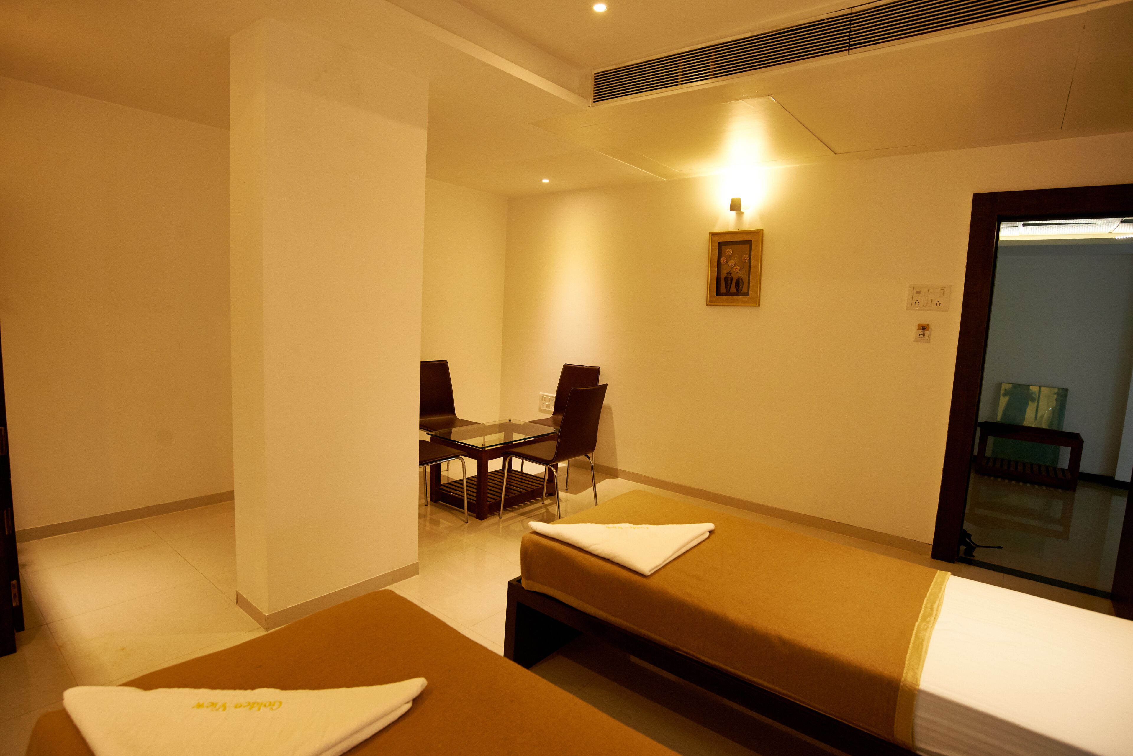 A hotel room with two beds, a small table with chairs, a painting on the wall, and a door leading to a bathroom.