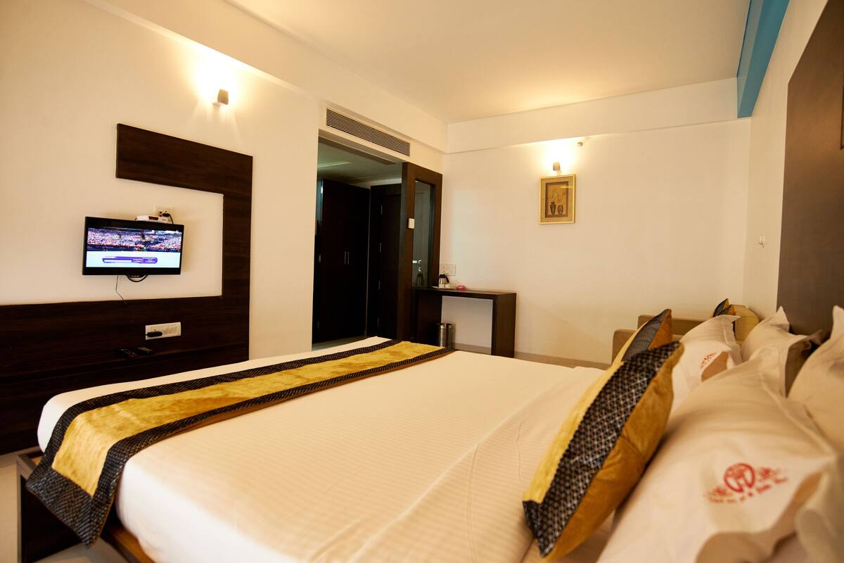 Habitación de hotel con una cama grande, un televisor fijado a la pared, un espejo y un escritorio pequeño con una lámpara.