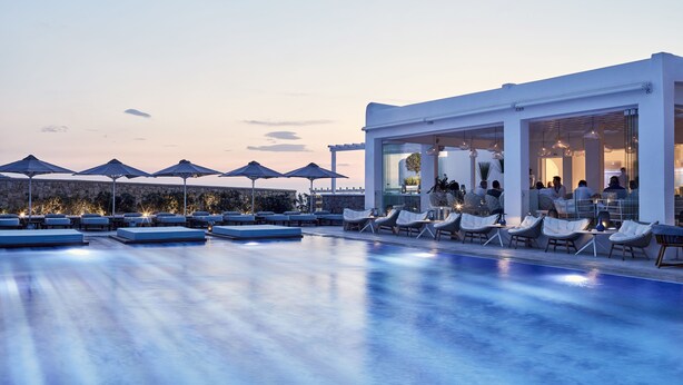 Un área de piscina moderna con sillones de descanso y sombrillas, un bar con paredes de vidrio y una vista despejada del cielo.