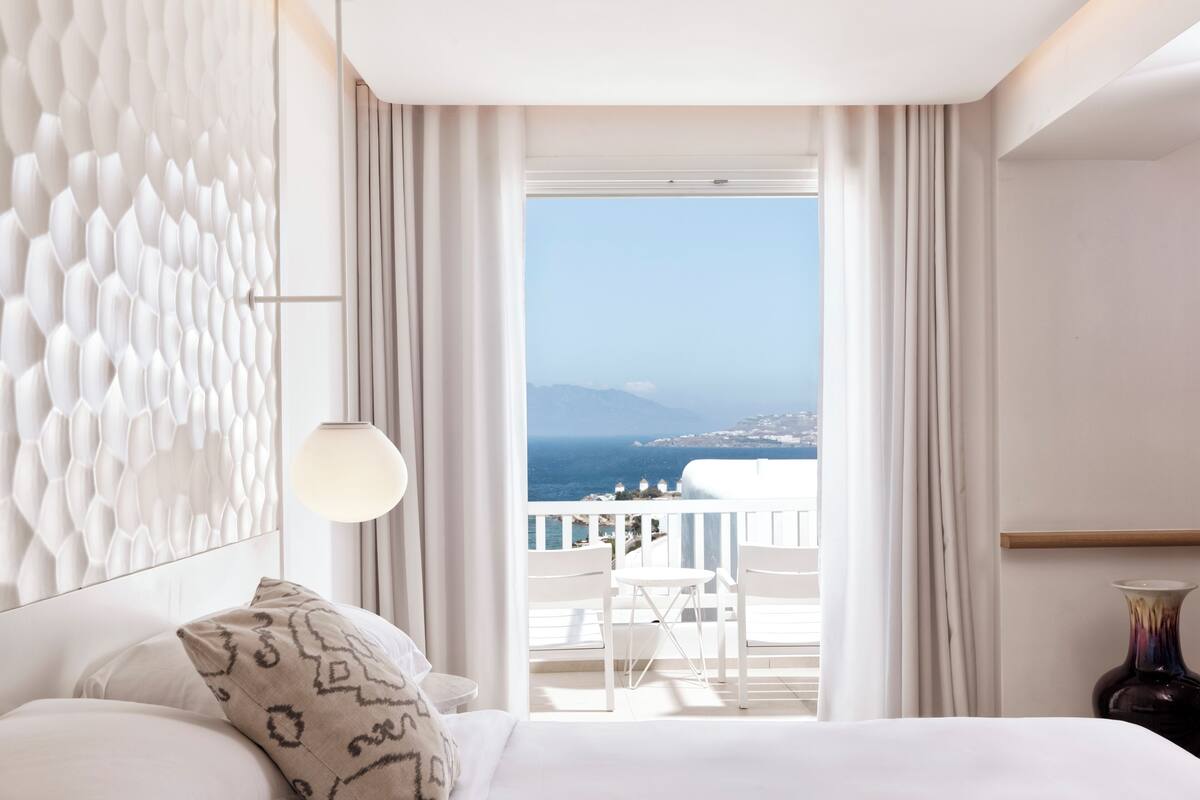 A hotel room with a bed, a patterned pillow, a wall with a textured design, a view of the sea and buildings through a window, and a vase on a side table.