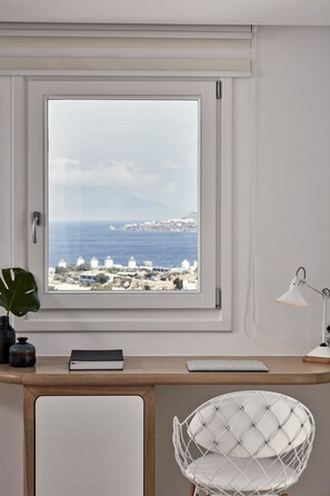 A room with a view of the sea and buildings, a desk with a chair, a lamp, and a plant.