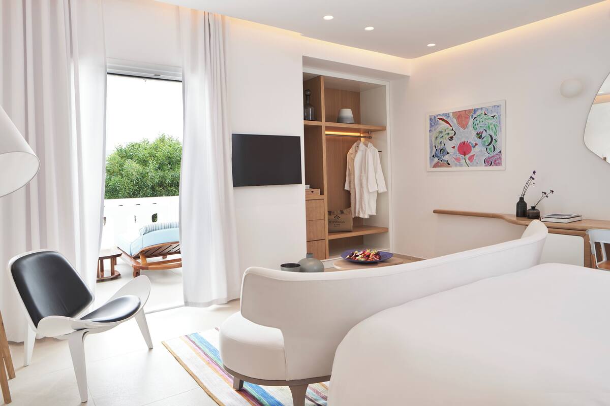 A modern hotel room with a bed, a seating area, a TV, a wardrobe, and a painting.