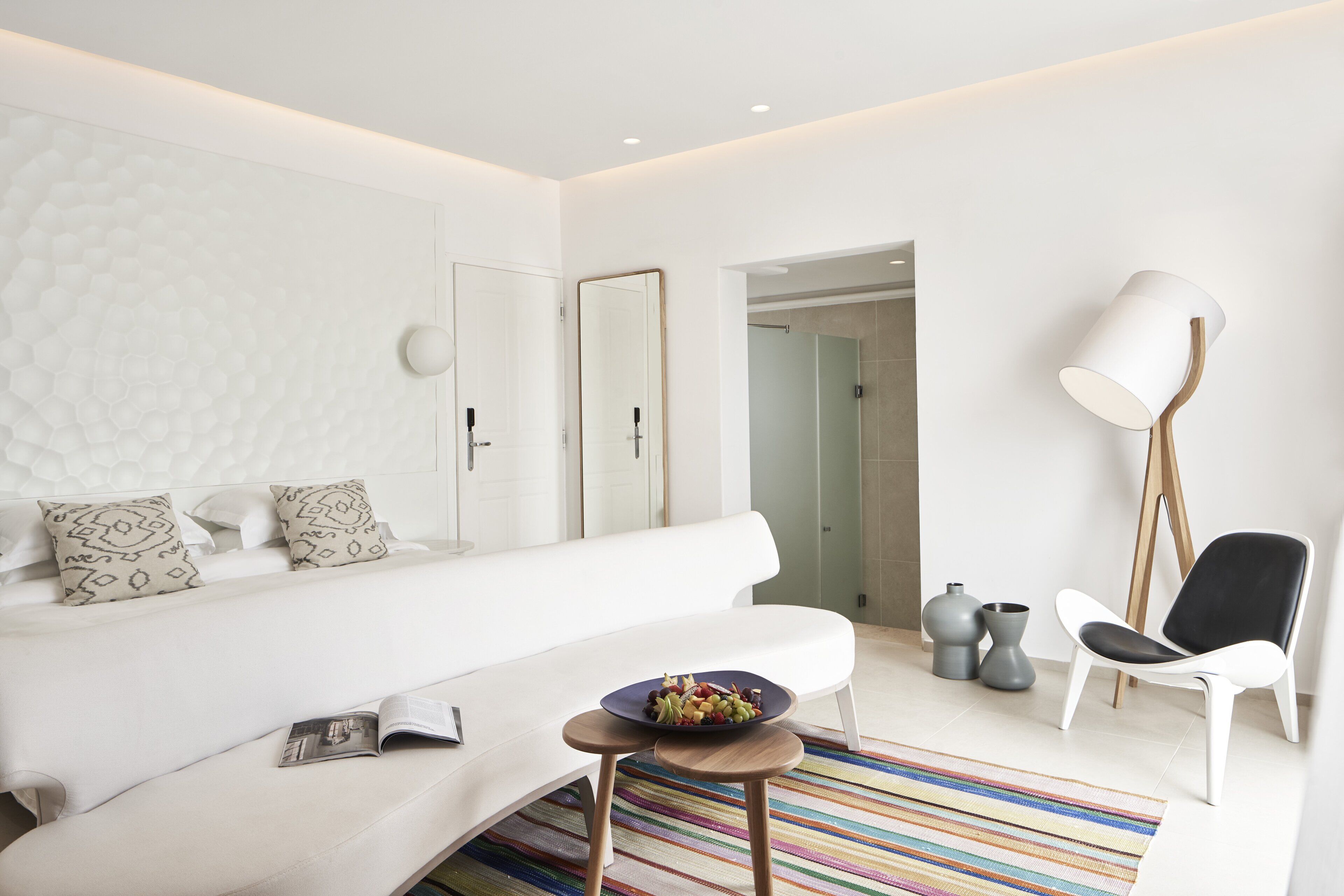 A modern, well-lit room with a white sofa, a small table with a bowl of fruit, a standing lamp, and a chair.