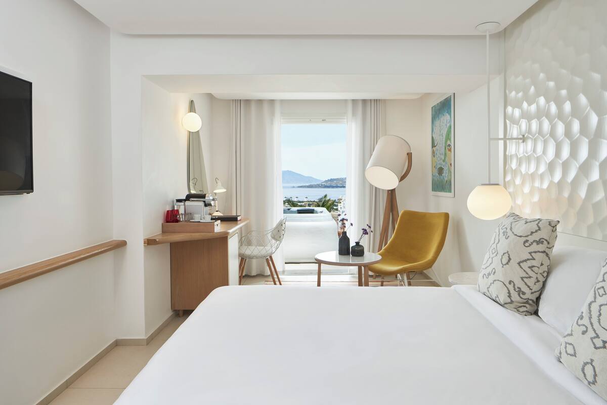 A hotel room with a bed, a desk, a chair, a table, and a view of the sea.
