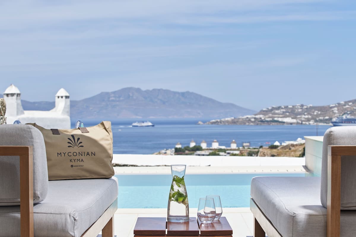 A poolside setting with a view of the sea and mountains, a Myconian Kyma bag, a glass vase with mint leaves, and two glasses on a wooden table.