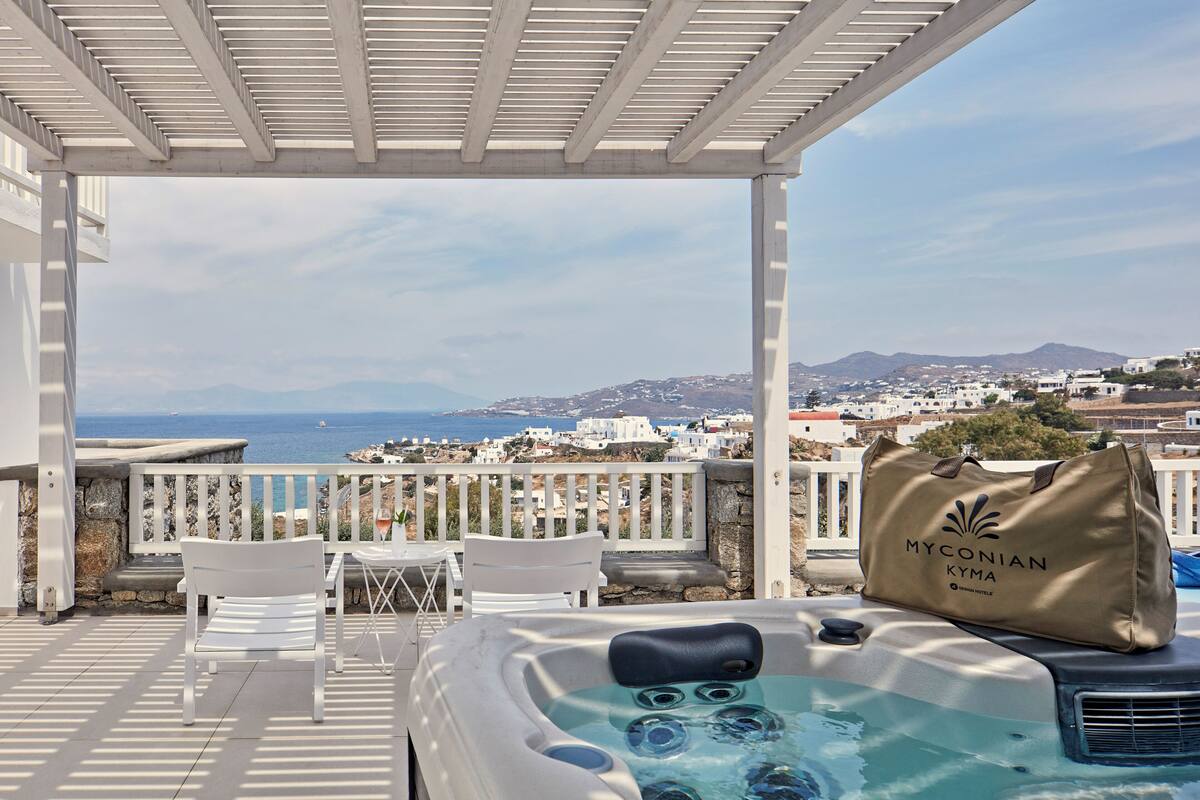A hot tub on a balcony with a view of the sea and a Myconian Kymata bag on the edge.