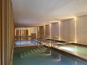 A modern indoor swimming pool with clear glass walls, metal railings, and a tiled floor.