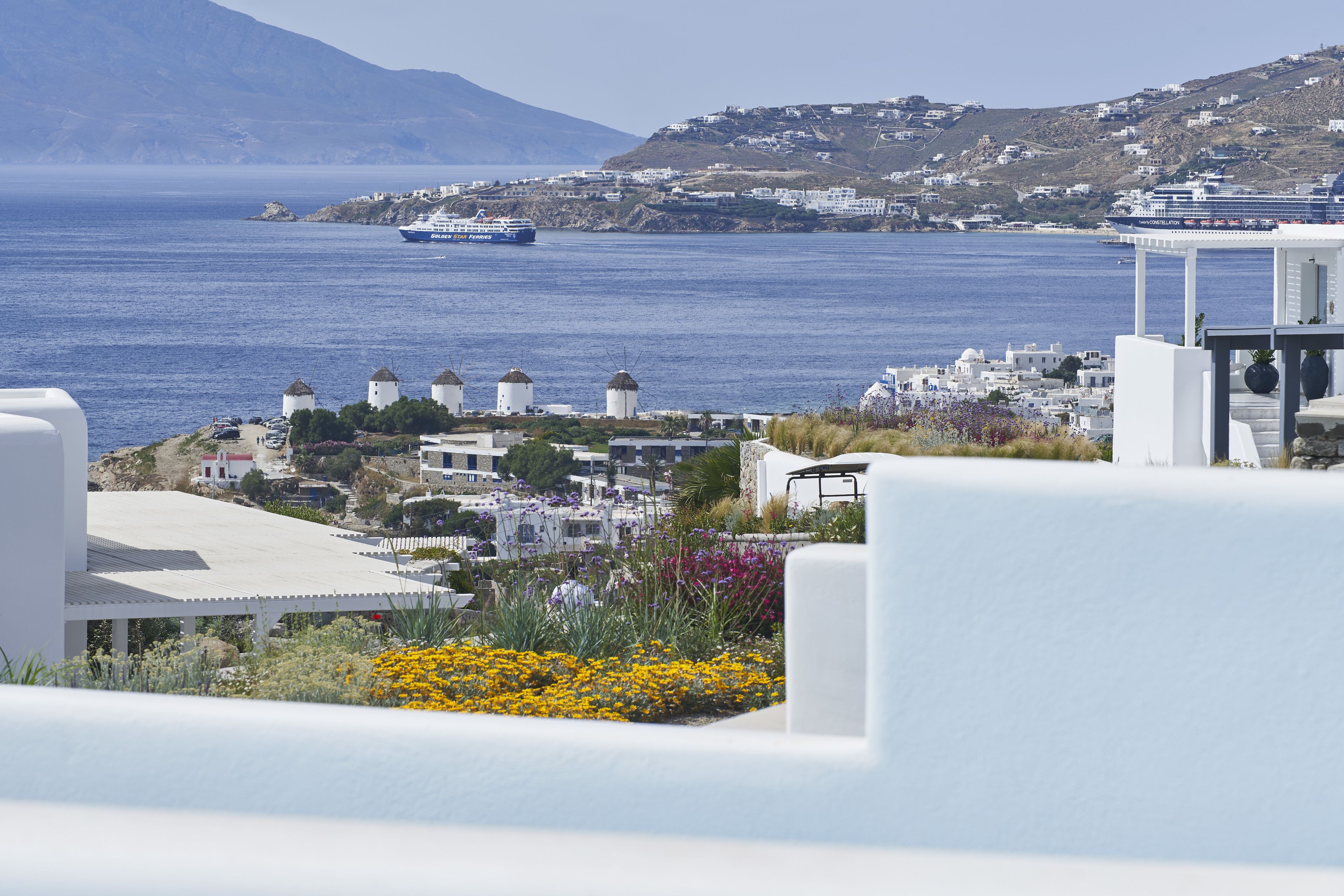 A coastal view with a white building, a ship in the distance, and a hillside with buildings.