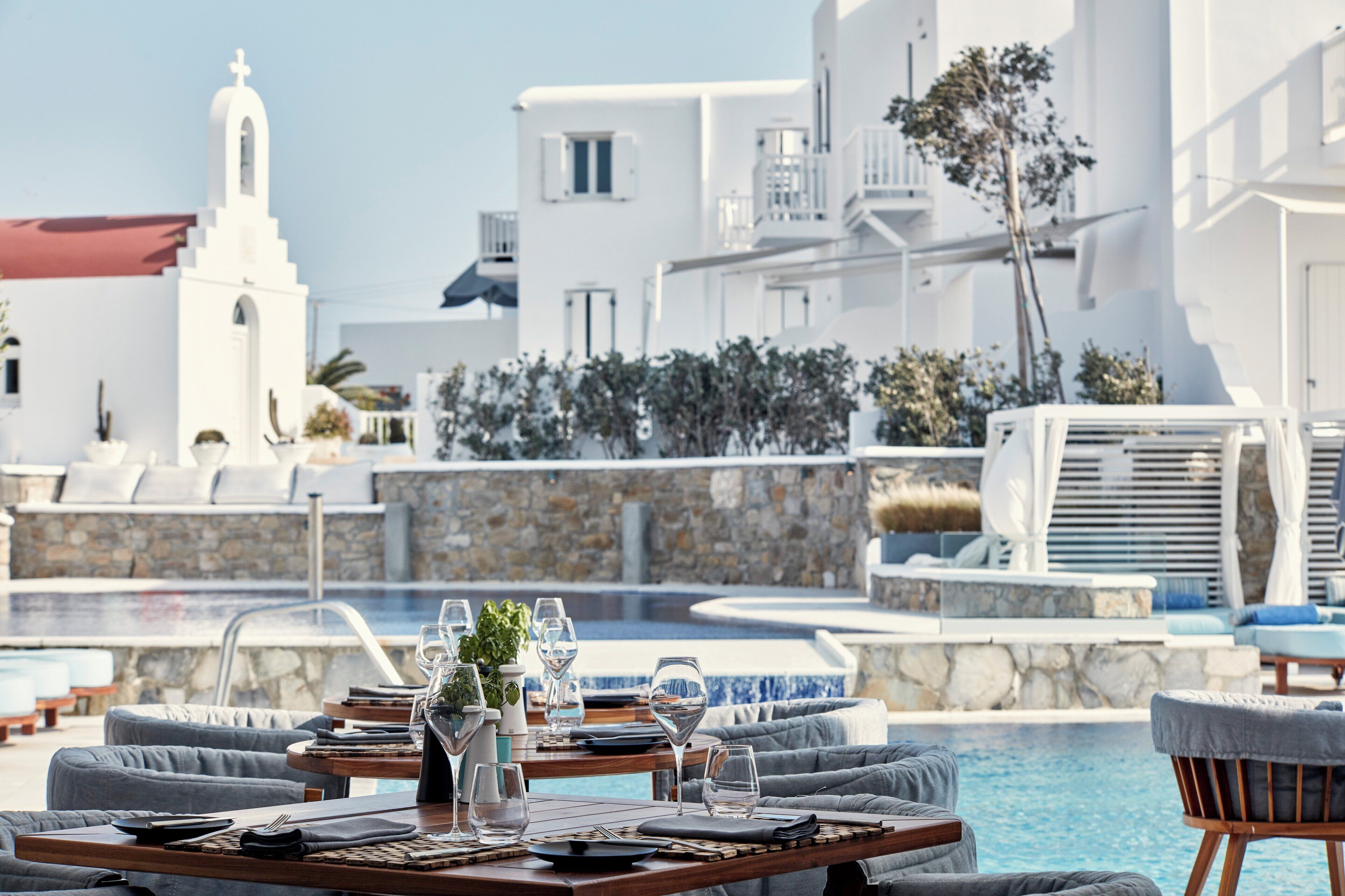A poolside dining area with tables set for two, a stone wall, and a white building with a bell tower in the background.