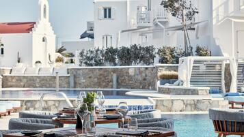 A poolside dining area with tables set for two, a stone wall, and a white building with a bell tower in the background.