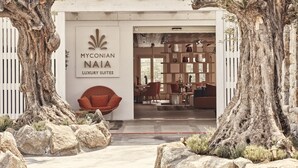 A modern building with a sign reading "MYCONIAN NAIA LUXURY SUITES". Inside, there are multiple seating areas with chairs and tables.