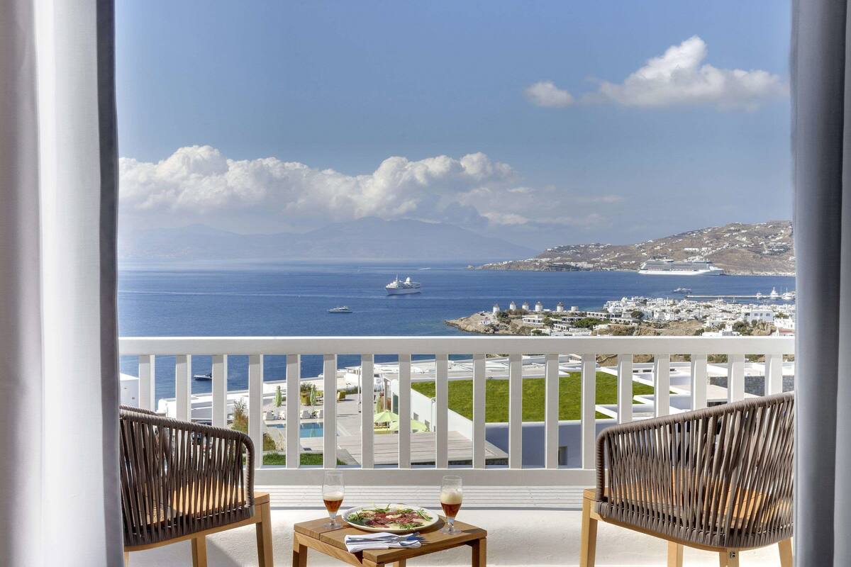 Un balcon agrémenté de fauteuils et d’une table en osier, offrant une vue dégagée sur une ville côtière, la mer et les bateaux.