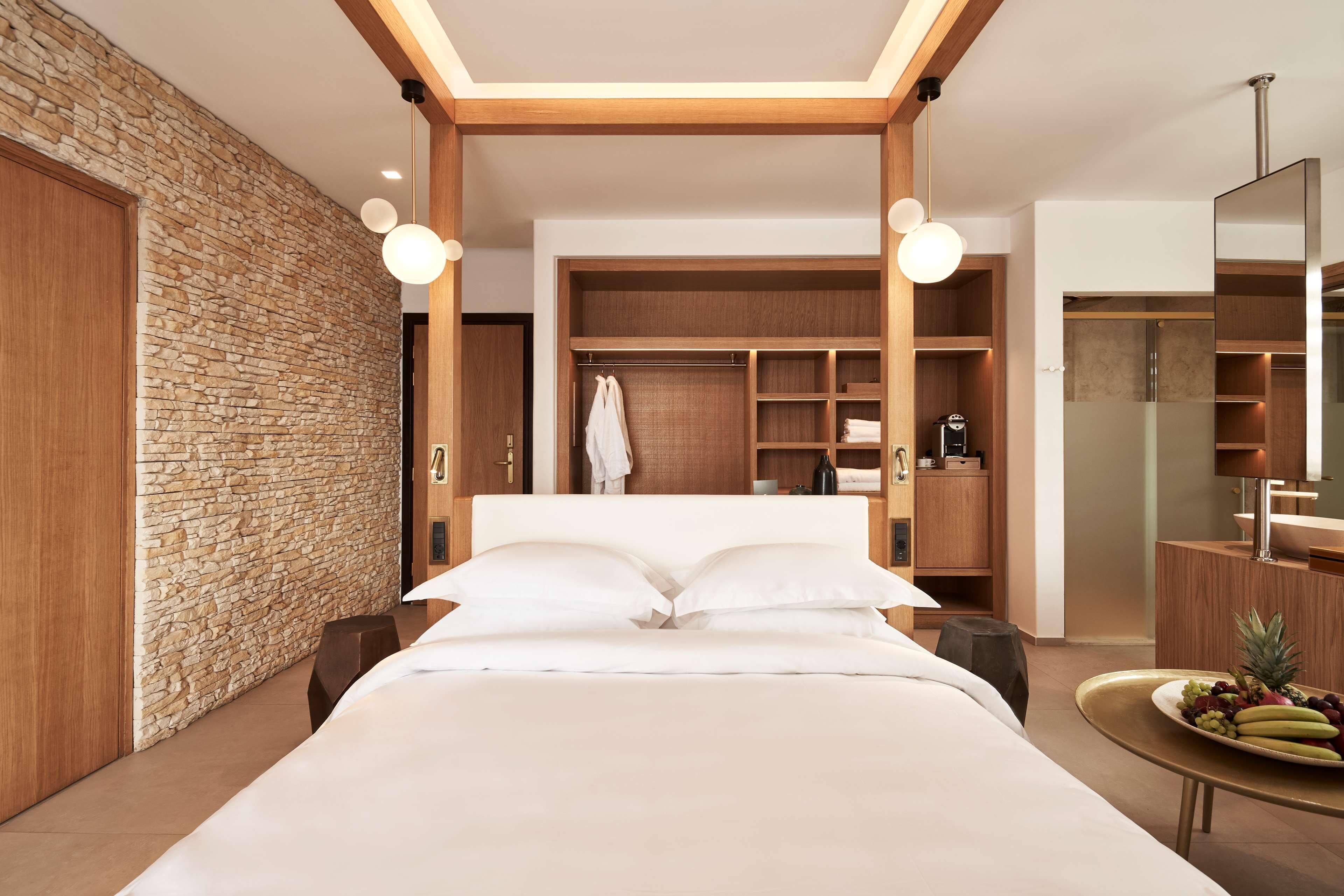 A modern hotel room with a stone accent wall, a large bed, built-in wardrobe, and a round table with fruit.