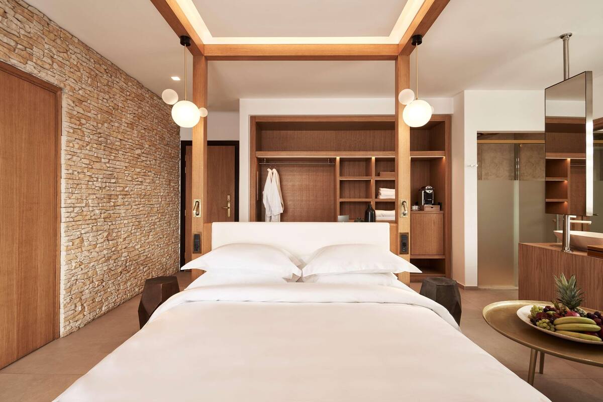 A modern hotel room with a stone accent wall, a large bed, built-in wardrobe, and a round table with fruit.