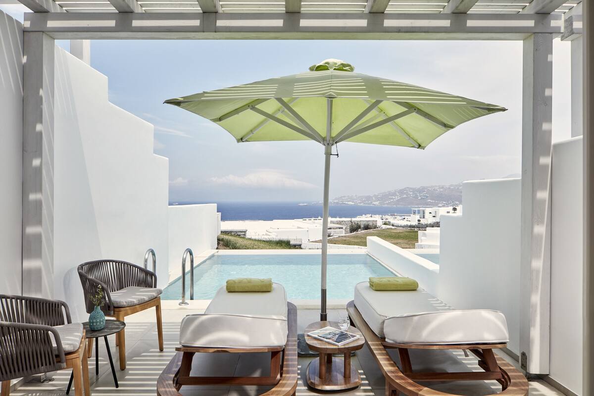 Une terrasse avec deux fauteuils, une table et un parasol, offrant une vue sur une piscine et la côte.