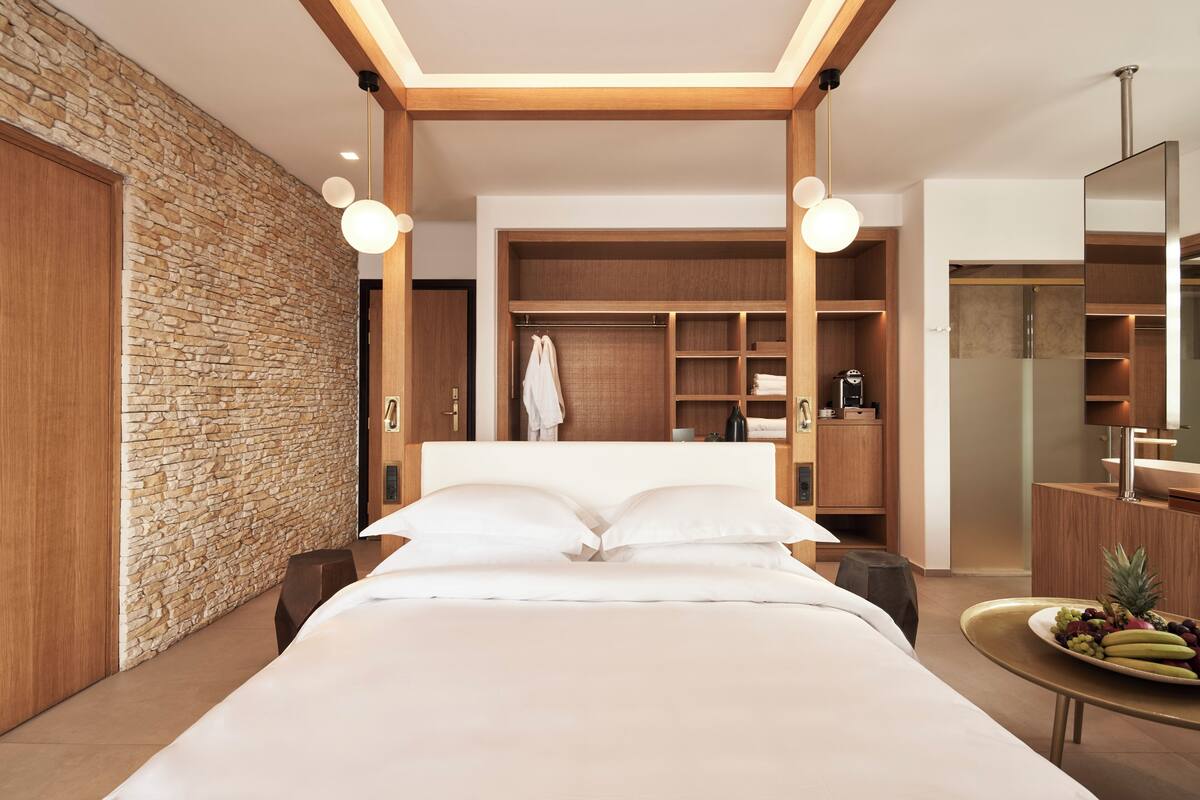 A modern hotel room with a stone accent wall, a large bed with white linens, a wooden wardrobe, a built-in vanity, and a fruit bowl on the side table.