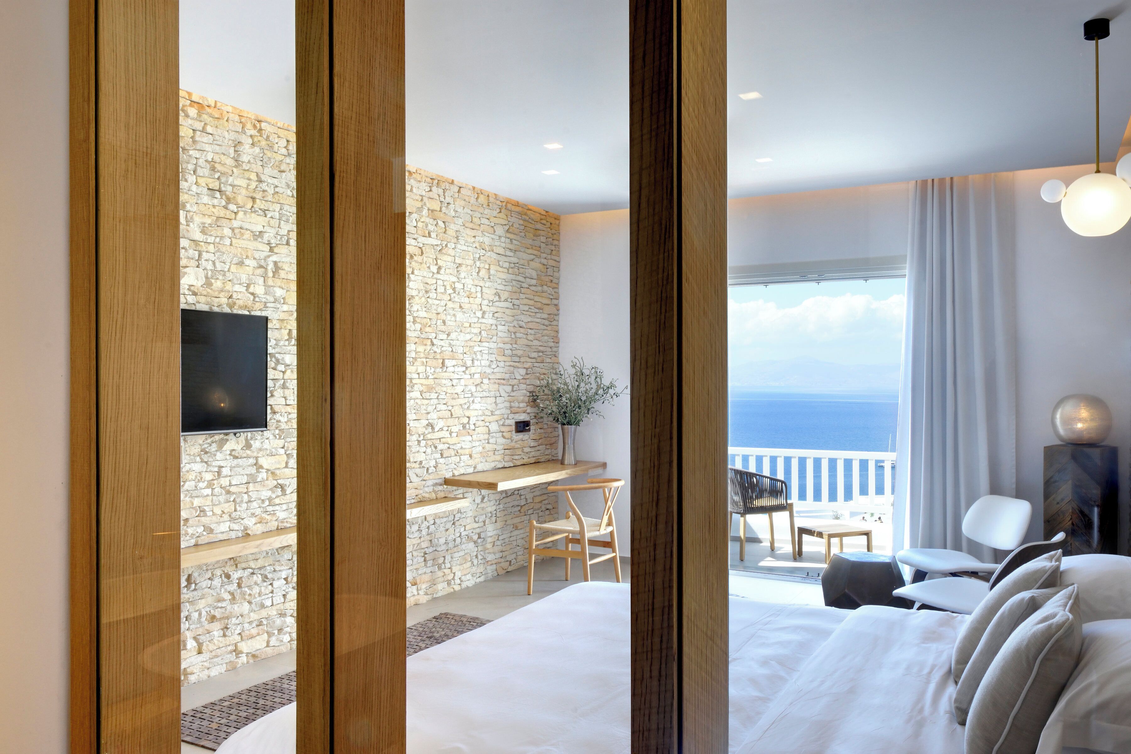A bedroom with a stone wall, a bed, a desk, and a view of the sea.