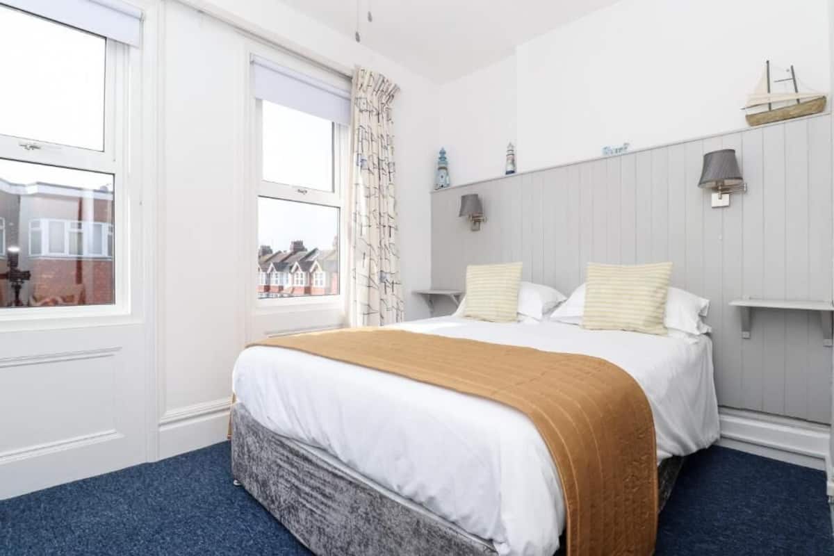 A hotel room with a large bed, a window with a view of houses, a curtain, and a wall-mounted lamp.