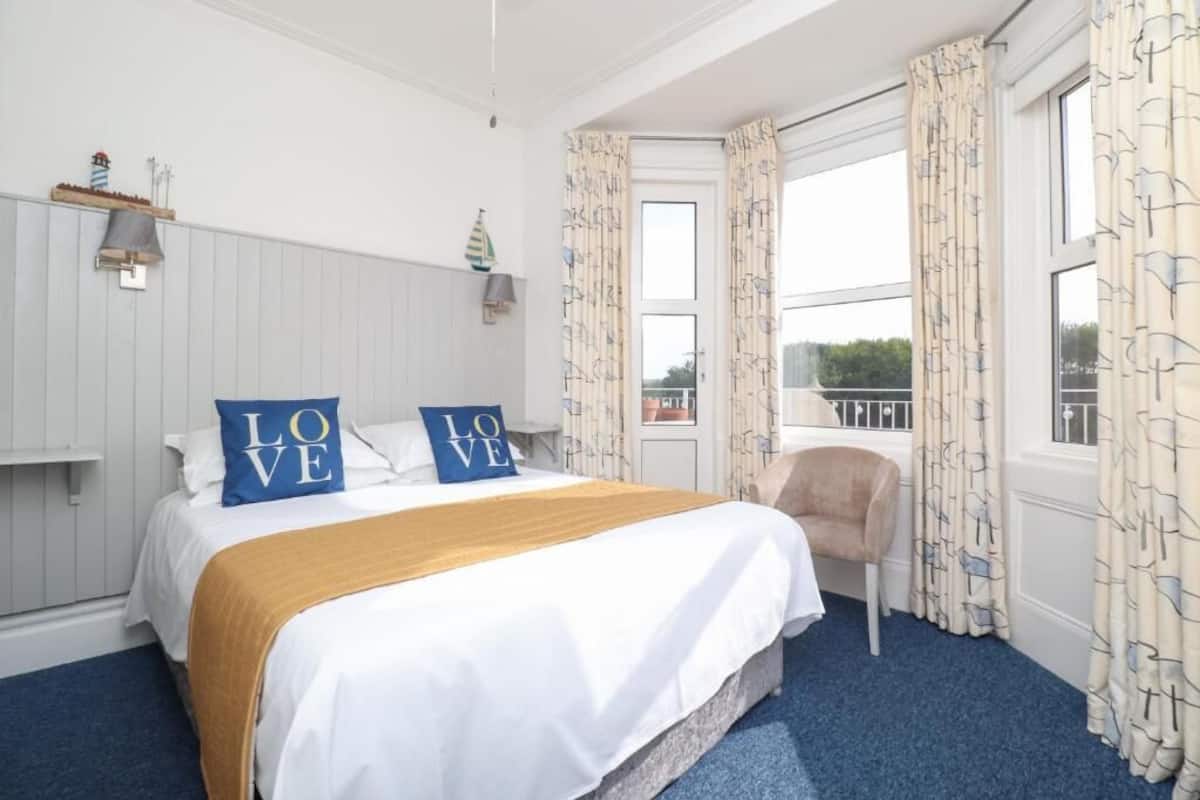 A hotel room with a bed, a chair, a window with curtains, a lamp, and a shelf with decorative items.