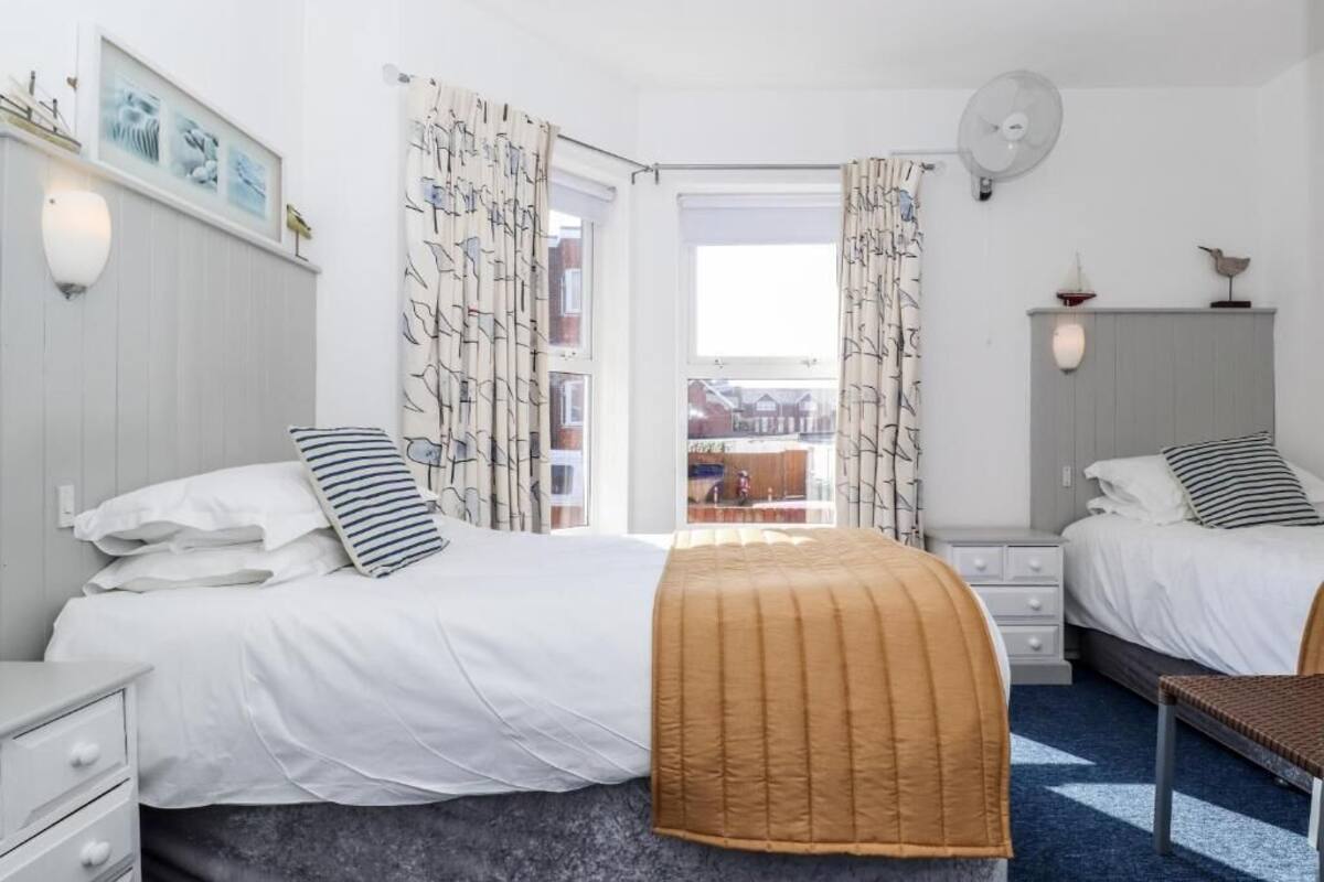 A bedroom with two beds, a window with curtains, a bedside table, a chair, and a fan.