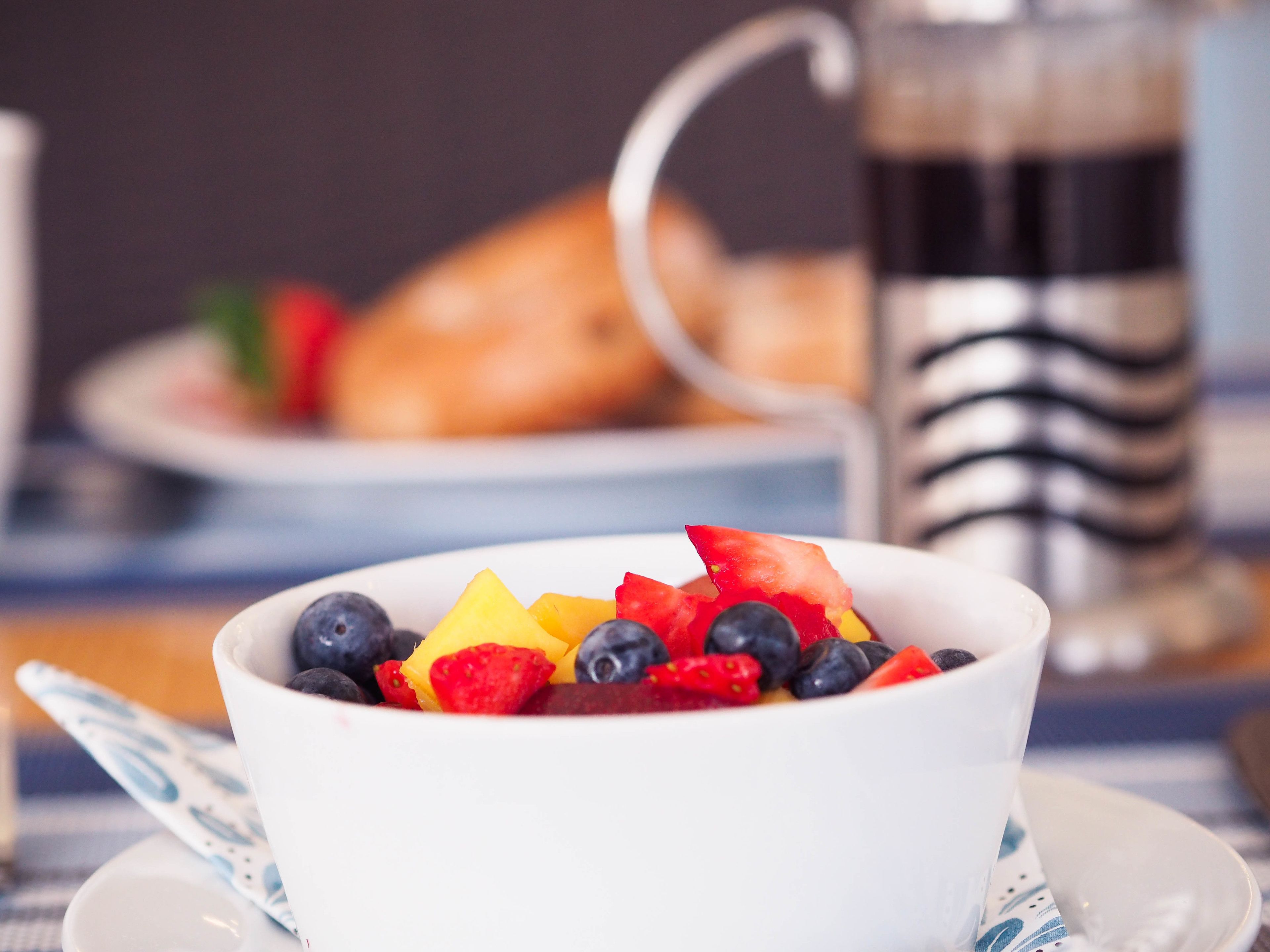 A bowl of fruit on a table with a cup and a pastry in the background.