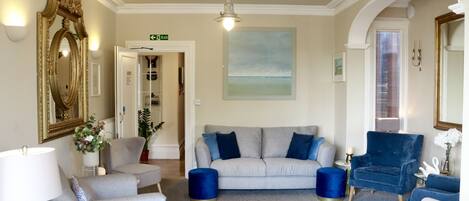 A spacious living room with a grey sofa, blue armchairs, and a white armchair. There is a large mirror, a painting, and a chandelier.