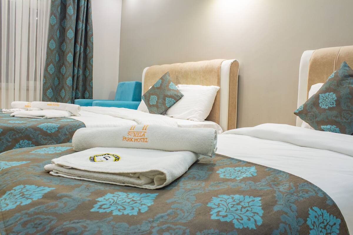 A hotel room with two beds, each with a patterned bedspread and pillows, a blue chair, and a window with sheer curtains.