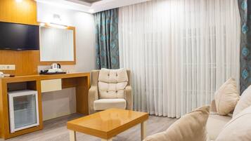 A modern hotel room with a wooden desk, a comfortable sofa, a cushioned armchair, a small refrigerator, and a flat-screen TV.