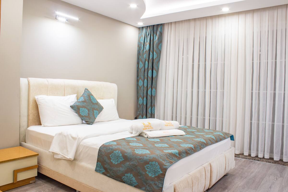 A neatly made bed with a patterned bedspread, a bedside table, and a window with sheer curtains.