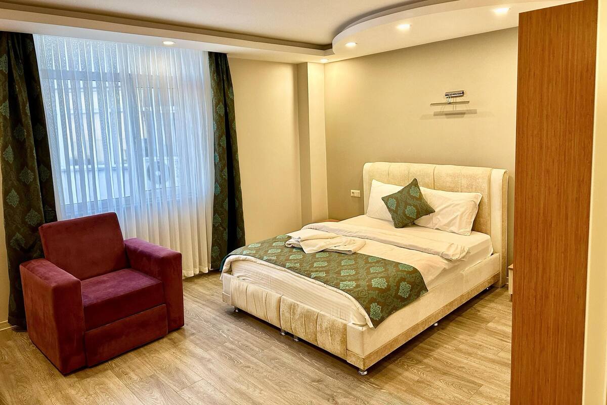 A hotel room with a bed, a red armchair, and a window with sheer curtains.