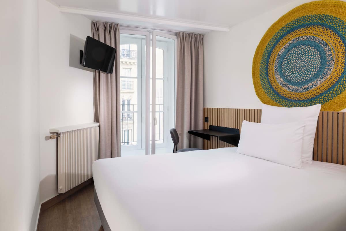 A hotel room with a bed, a TV mounted on the wall, a window with curtains, a view of the outside, and a wooden headboard.