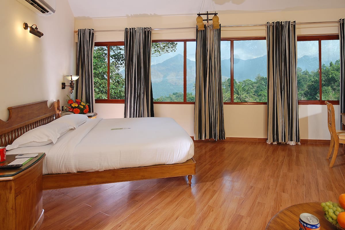 Habitación de hotel con cama, escritorio, silla y vistas a montañas a través de amplios ventanales.
