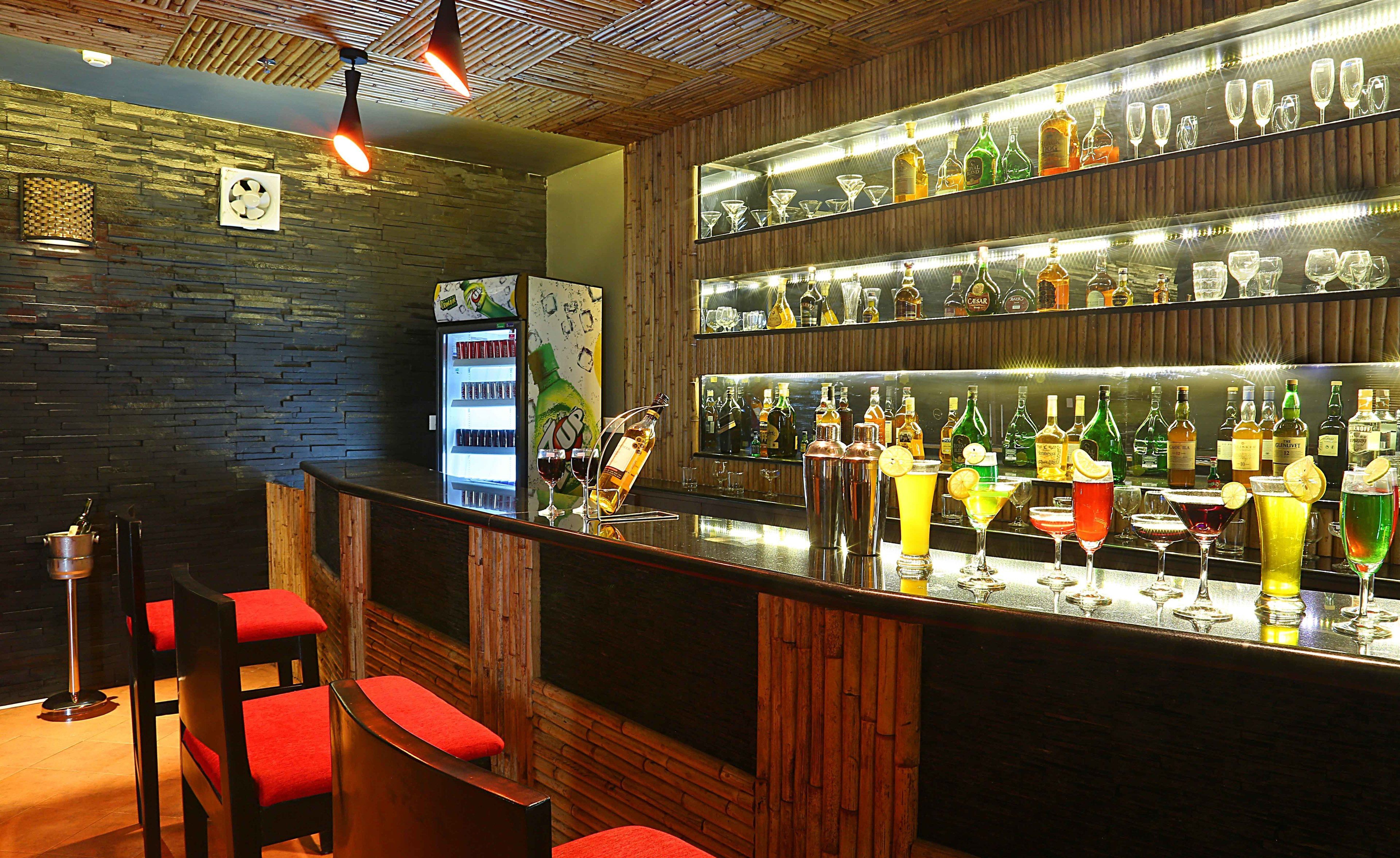 Una zona de bar con mostrador de madera, taburetes con cojines rojos y estanterías llenas de botellas de licor y vasos.