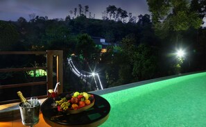 Una mesa con frutero, botella de champán y copa de bebida roja, con vista a un puente iluminado y una ladera boscosa.