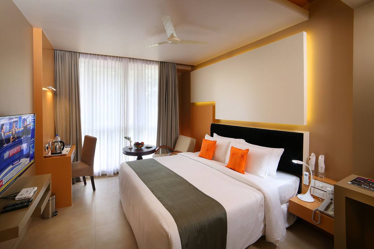 Habitación de hotel con cama, escritorio con televisor, silla, mesita con un florero y ventilador de techo.