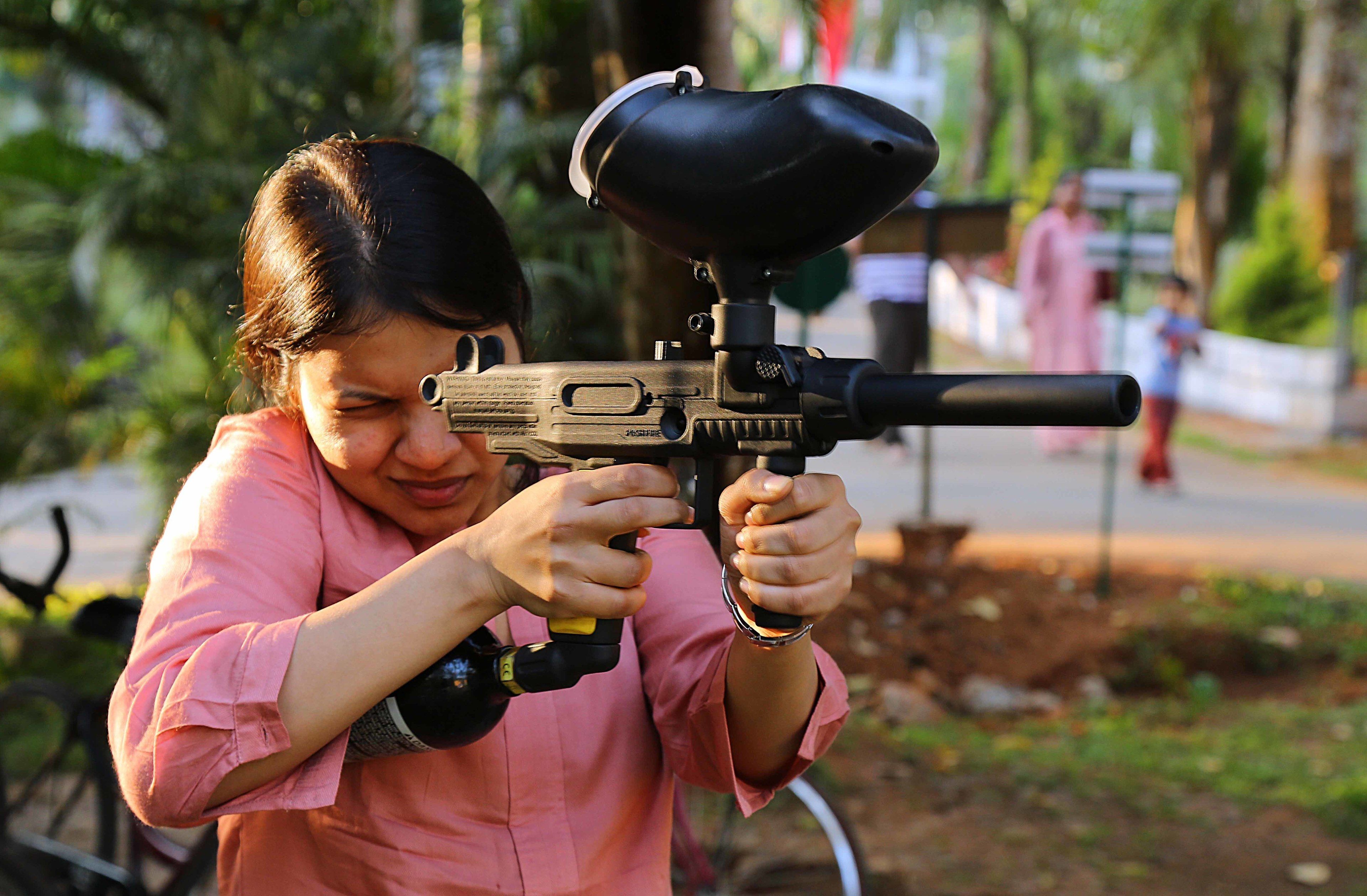 A person holding a toy gun with a camera attached, in an outdoor setting with greenery and a pathway.
